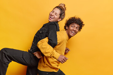 Positive female and male friends foolishing around gve piggyback ride to each other laugh happily dressed in black and yellow clothes pose indoor. Girlfriend and boyfriend spend free time together