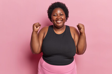 Positive overweight African American woman clenches fists and smiles broadly roots for favorite...