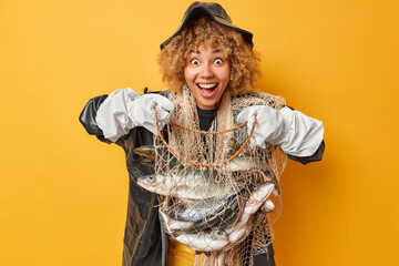 Positive glad female fisher wears black hat raincoat gloves holds net full of variety of fish has...