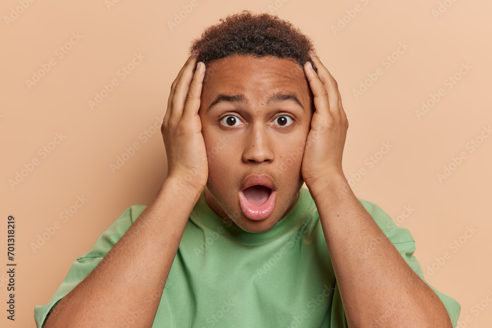 Wall mural People emotions concept. Indoor photo of young confused African american guy holding head with hands looking straight at camera standing in centre isolated on beige background not knowing what to do - Wall murals