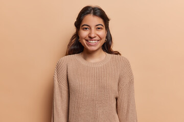 Portrait of pleasant looking Iranian girl with toothy smile has good mood dressed in casual knitted jumper looks directly at camera isolated over brown background. People and positive emotions concept - obrazy, fototapety, plakaty