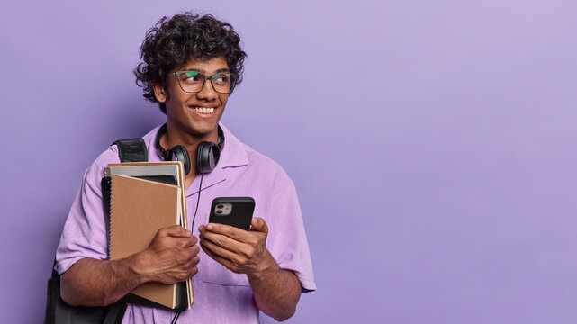 Student lifestyle. Indoor waist up of young happy smiling broadly Hindu guy wearing purple shirt standing on left isolated holding books and smartphone looking at blank space for your advertisement