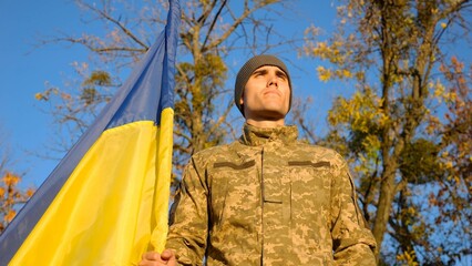Ukrainian army man stands with national banner at countryside against blue sky. Male soldier in military uniform with Ukraine flag as symbol of victory against russian aggression. Invasion resistance.