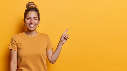 Beautiful young European woman with hair bun smiles pleasantly points index finger on copy space...
