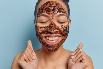 Positive young woman applies coffee mask for cleansing skin keeps eyes closeed and smiles toothily...