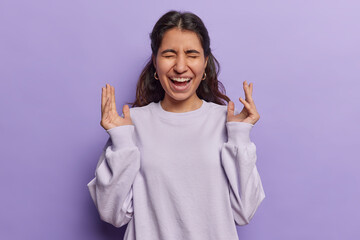 Waist up shot of overjoyed Iranian woman with dark hair exclaims loudly keeps hands raised and...