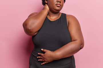 Cropped shot of unrecognizable dark skinned woman dressed in black sportswear touches neck and stomach goes in for sport to loose weight poses against pink background . Faceless body positive female