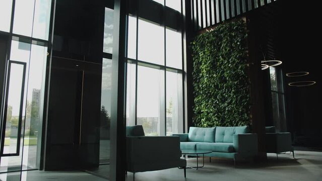 Dolly shot of empty hall waiting place, shadows on dark turquoise sofa, sofa near the window in the hall of the building greenery wall background, slow motion dolly shot.