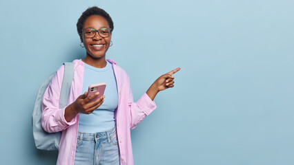 Horizontal shot of cheerful dark skinned woman dressed in casual pink shirt and jeans holds mobile...