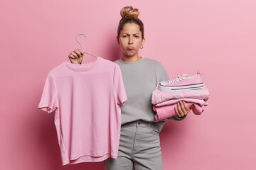 Female shopaholic feels frustrated looks unhappily holds neatly folded clothing and tshirt on...