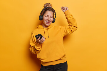 Carefree joyful young woman enjoying awesome beat in new stereo headphones shakes arms and exclaims loudly closes eyes from pleasure dressed in casual hoodie isolated over vivid yellow background. - obrazy, fototapety, plakaty