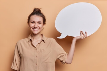 Idea and inspiration. Positive smiling woman with glad expression wears casual shirt holds blank speech bubble for your advertising content isolated over brown background. Mock up. Amazing offer