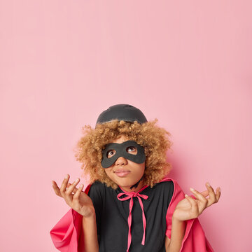 Vertical shot of hesitant female superhero spreads palms and concentrated above with clueless expression wears helmet helmet eyemask and cloak isolated over pink background copy space for your promo