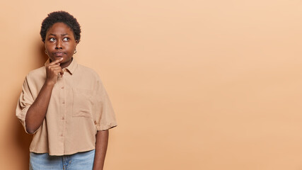 Horizontal shot of thoughtful dark skinned African woman keeps hand on chin looks aside pensively wears shirt and jeans isolated over brown background copy space for your advertising content