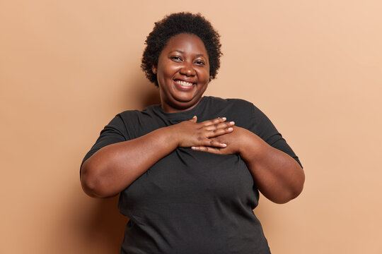 Kindness appreciation and gratitude concept. Smiling dark skinned overweight young African woman presses heart being thankful dressed in casual black t shirt isolated over beige studio background.