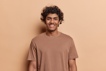 Portrait of happy Hindu man with curly hair smile toothily keeps arms down dressed in casual t shirt looks cheerful isolated over brown background being in good mood. Human positive emotions concept
