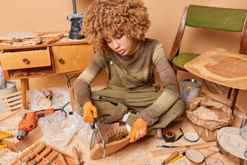 Indoor shot of curly haired female carpenter cuts pieces of furniture wears jumper and overalls...