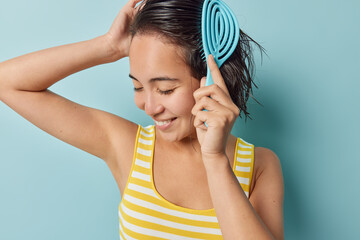 Photo of cheerful dark haired young Asian woman combs wet hair after showering smiles gladfully keeps eyes closed wears yellow striped t shirt isolated over blue background. Hair care concept - obrazy, fototapety, plakaty