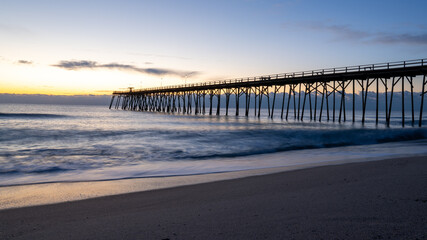 Sunrise casts a warm glow over Kure Beach pier, with soft waves lapping the shore in North Carolina.