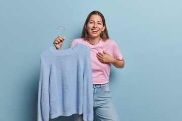 Photo of overjoyed young European woman laughs happily holds knitted sweater on hanger suggests to...