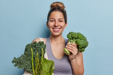 Sporty young woman in grey top holds green vegetables keeps to healthy nutrition smiles broadly...