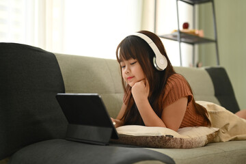 Little Asian girl in headphone lying on couch and watching video on digital tablet.