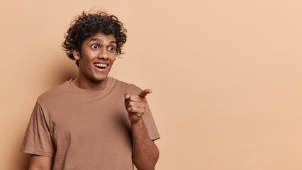 Positive surprised curly haired adult Hindu man looks with great surprise aside points index finger...