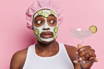 Shocked African man with beard applies nourishing clay mask and cucumber slices for skin...