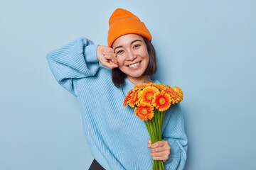 Asian woman revels in holiday joy holds bouquet of orange daisy flowers expresses happy emotions wears hat and knitted jumper isolated over blue background. People celebration and pure happiness