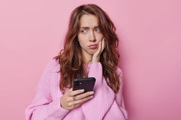 Horizontal shot of upset long haired woman holds smartphone ponders on how to solve problem holds smartphone dressed in casual jumper poses against pink background. Pensive female model with gadget