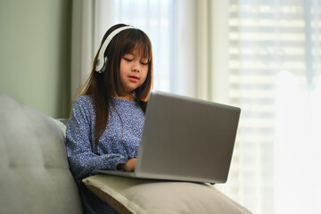 Cute schoolgirl in headphone watching online virtual class on laptop. Education concept.
