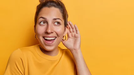 Fotobehang Horizontal shot of healthy young cheerful woman keeps hand near ear tries to overhear rumors listens private information with interest has curious expression isolated over yellow background. © Wayhome Studio