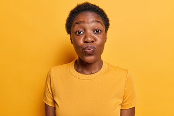 Human facial expressions. Lovely dark skinned woman keeps lips folded focused at camera has romantic mood dressed in casual t shirt isolated over vivid yellow background wants to kiss someone