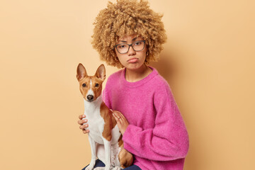 Upset curly woman looks sadly at camera purses lips poses with pedigree dog spends free time with best friend sits on chair has adorable pet wears spectacles and pink sweater isolated on beige wall