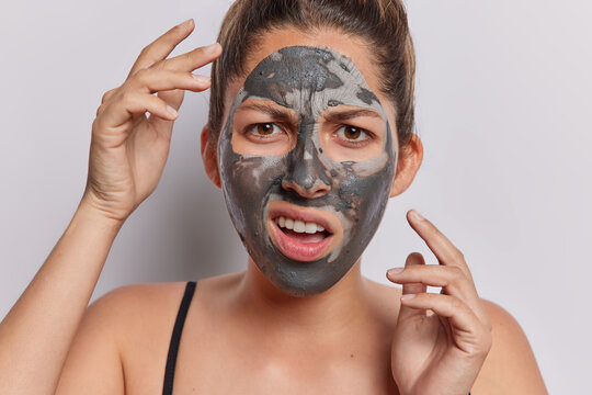 Photo of displeased young woman frowns face from dissatisfaction applies clay mask on face for skin rejuvenation stands bare shouldered indoor against white background. Beauty procedures concept
