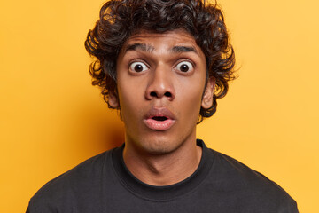 Portrait of stupefied young Hindu man with curly hair stares bugged eyes at camera holds breath and...