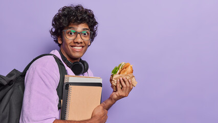 People positive emotions concept and student lifestyle. Studio sideway photo of young happy smiling Hindu male holding books and burger standing on left isolated on purple background with space