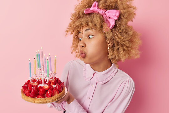 People and celebration. Indoor sideway photo of young pretty African american female having party standing in centre isolated on pink background holding cake with strawberries blowing candles