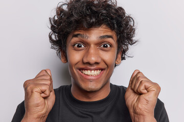 Thrilled positive handsome curly haired Hindu man clenches fists smiles broadly supports someone...