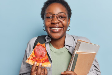 Cheerful dark skinned teenage girl being student at university eats pizza during break poses with...