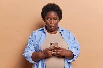 Photo of impressed dark skinned African woman stares in smartphone display being deeply surprised...