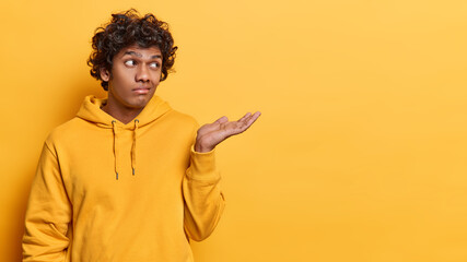 Confused hesitant Hindu man shrugs shoulder keeps palm raised up dressed in casual sweatshirt doesnt know why happened like this isolated over yellow background copy space for your promotion - obrazy, fototapety, plakaty