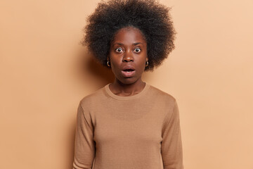 Fototapeta na wymiar Waist up shot of shocked African woman with dark bushy hair has scared horrified expression dressed in casual clothing holds breath from amazement isolated over brown background. Omg concept
