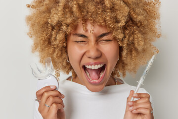 Horizontal shot of emotional curly haired European woman holds syringe and mouth expander exclaims...