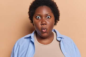 Portrait of shocked dark skinned overweight woman stares bugged eyes keeps lips rounded cannot believes in something very terrible dressed in blue shirt isolated over brown background. Omg concept