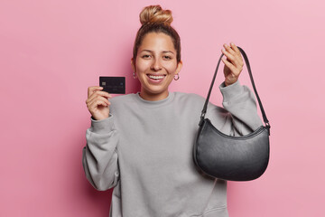 Waist up shot of positive European woman consumer holds new leather bag and credit card makes...