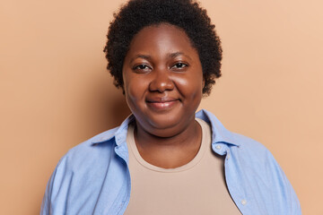 Portrait of pleasant looking curly haired chubby young African woman with curly hair dressed casually poses satisfied against brown background makes photo. People ethnicity and emotions concept