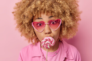 Headshot of surprised blonde curly haired European woman covers mouth with round lollipop has sweet tooth wears sunglasses dressed in jacket isolated over pink background. Harmful food concept