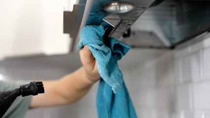 Woman wiping exhaust hood in kitchen, closeup. A woman's hand washes the hood in the kitchen with a rag. Cleaning a household hood. Female hands cleaning a kitchen hood with a cloth. - obrazy, fototapety, plakaty