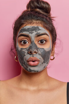 Studio photo of young beautiful surprised European woman standing in centre isolated on pink background applying face clay mask to make skin fresh looking straight at camera keeping hair in bun
